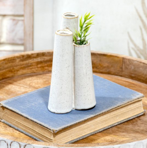 Triple bud white ceramic vase with a plant on top of a blue book on a wooden surface.