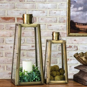 Two gold lanterns with candles on a wooden surface against a brick wall.