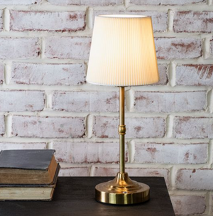 Gold table lamp with a white shade on a wooden surface against a white brick wall.